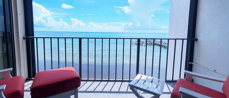 Un balcon avec deux fauteuils rouges dotés de coussins et une table blanche, d’où l’on peut admirer une mer bleue et limpide ainsi qu’une jetée.