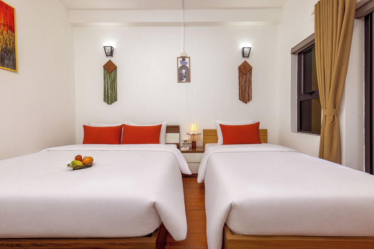 A hotel room with two beds, a fruit basket, wall-mounted lamps, and a window with curtains.