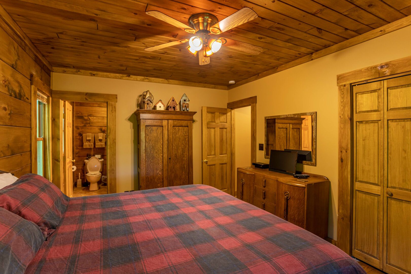 A bedroom with a bed, wooden ceiling, a ceiling fan, a television on a wooden cabinet, and a wooden door.
