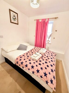 A bedroom with a bed, a window with pink curtains, a wall-mounted picture, and a ceiling light fixture.