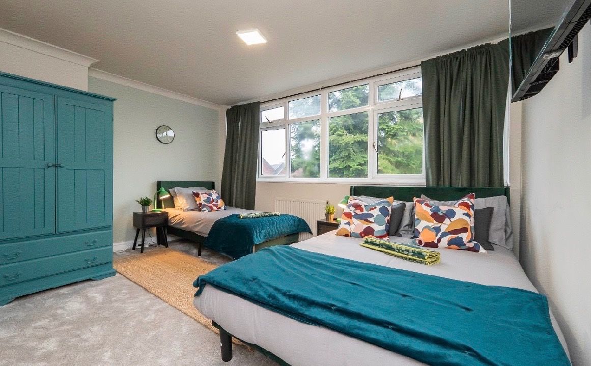 A bedroom with two beds, a wardrobe, a window with curtains, and a bedside table with a lamp.