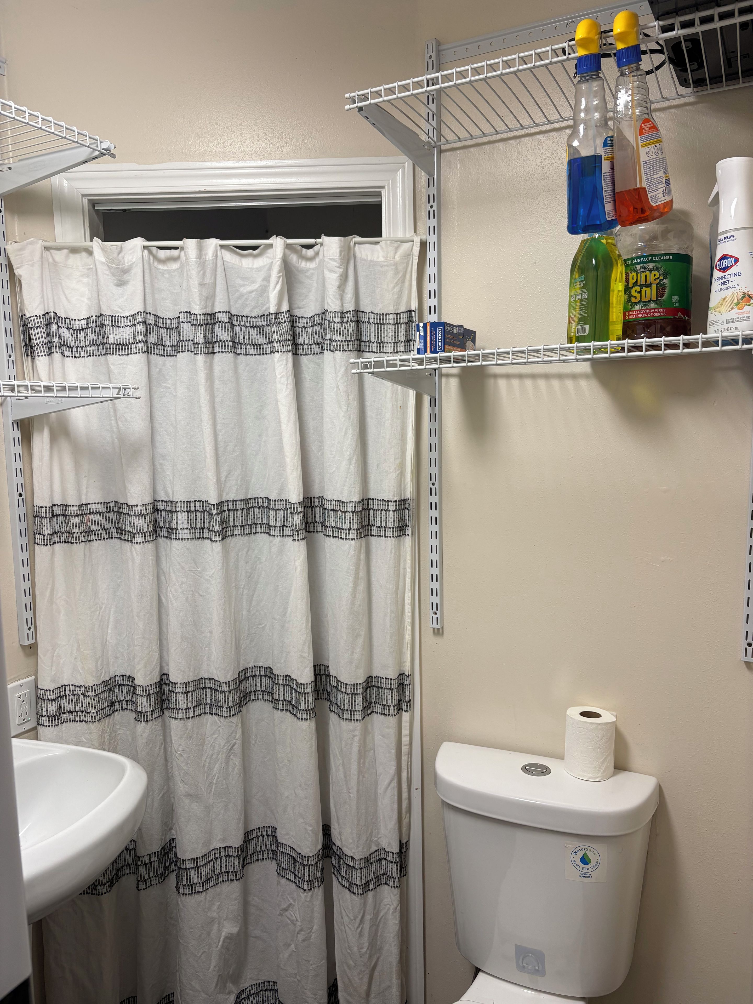 A bathroom with a shower curtain, a toilet, and a shelf with cleaning supplies.