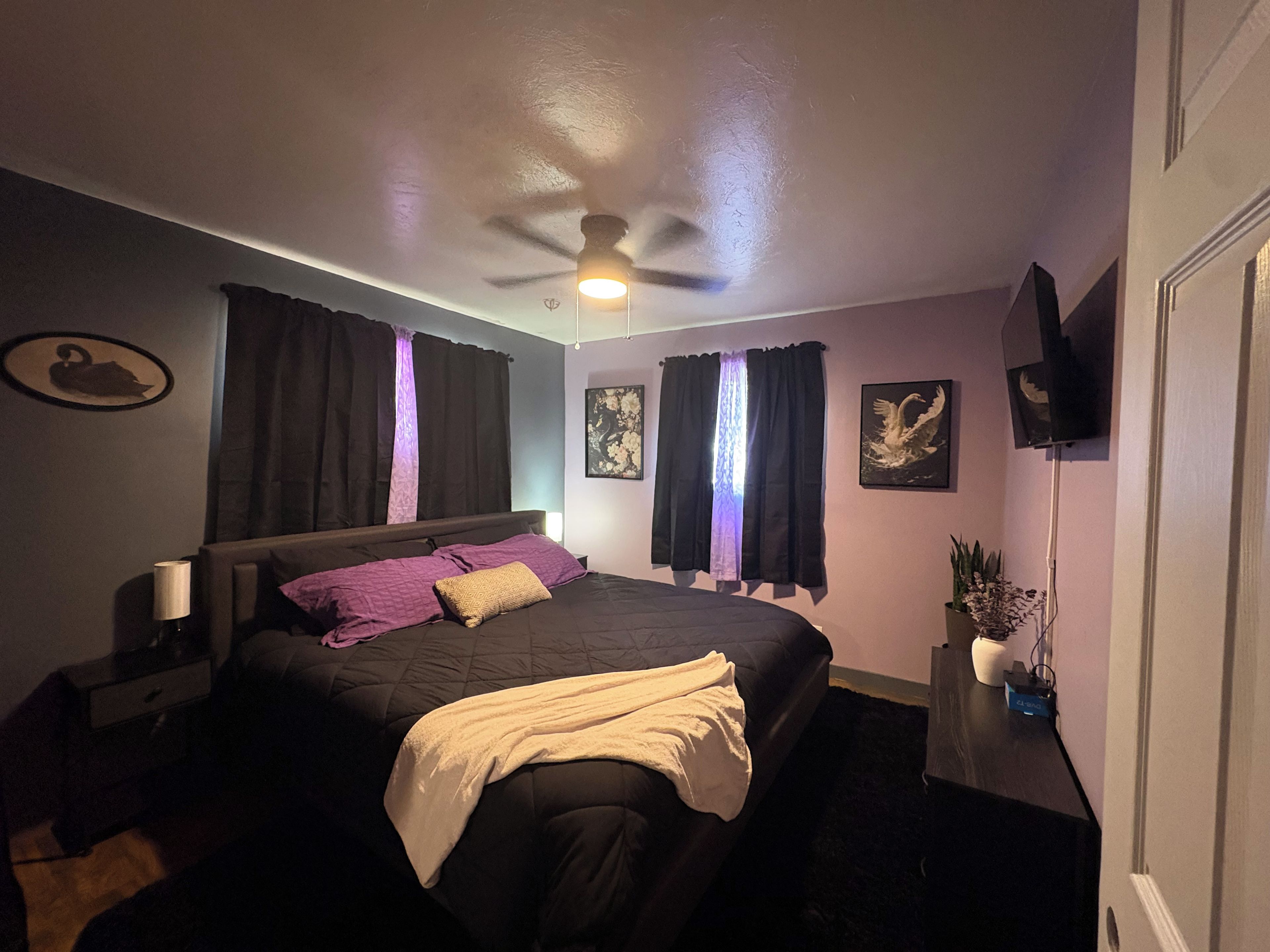 Un dormitorio con cama, ventilador de techo, mesita de noche, televisor y una pared decorada con obras de arte.