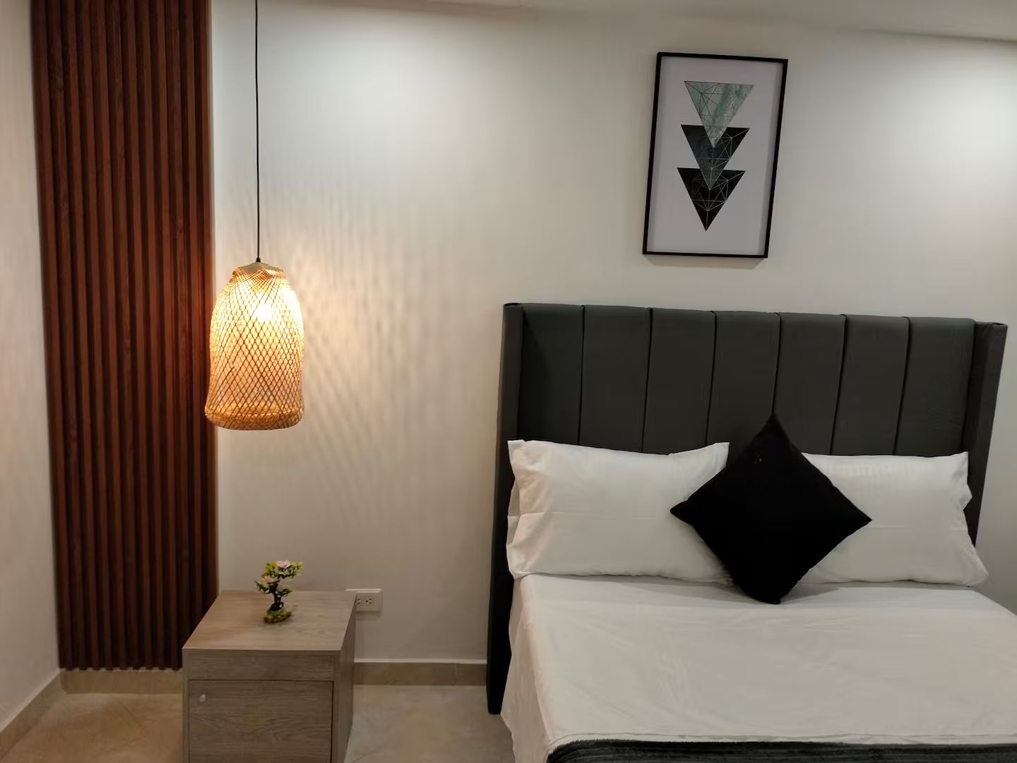 A neatly arranged hotel room with a bed, a bedside table with a vase of flowers, a hanging light fixture, and a framed geometric artwork on the wall.