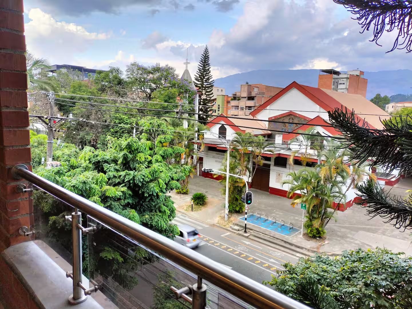 A view from a balcony overlooking a street with buildings, including a church with a cross on top, and lush greenery.