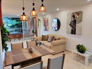 A modern living room with a sofa, dining table, and chairs. There are hanging lights, a painting, and a view of the outdoors. - Exclusive, luxurious, centrally located apartment — serene and comfortable 302 (Medellín)
