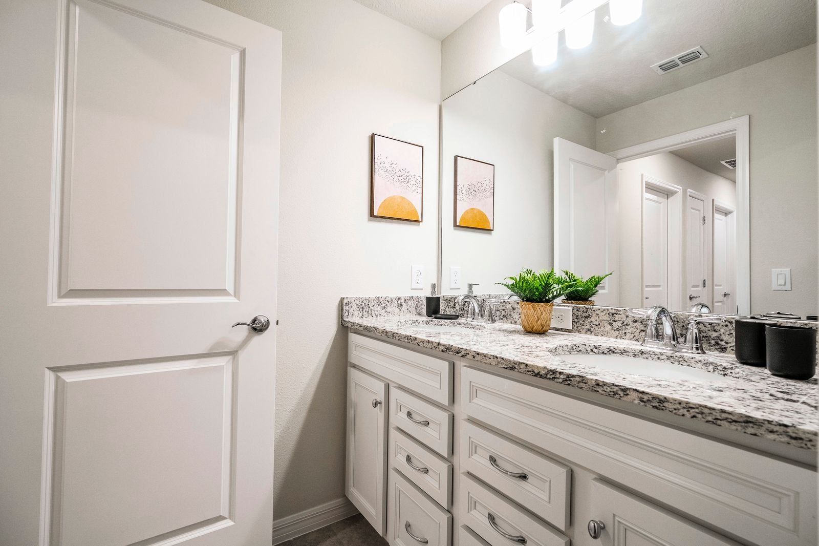 A bathroom with a white door, a granite countertop, a sink with a mirror, and two framed pictures on the wall.