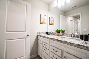 Un baño con puerta blanca, encimera de granito, lavabo con espejo y dos cuadros en la pared.