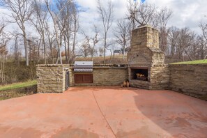 An outdoor stone barbecue area with a grill and a wood-fired oven, surrounded by a stone wall and trees.