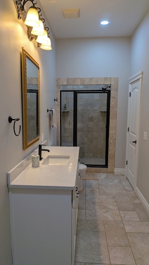A bathroom with a white vanity, a large mirror, a shower with a glass door, and a light fixture above the mirror. - Hillside Hideaway (Bentonville)