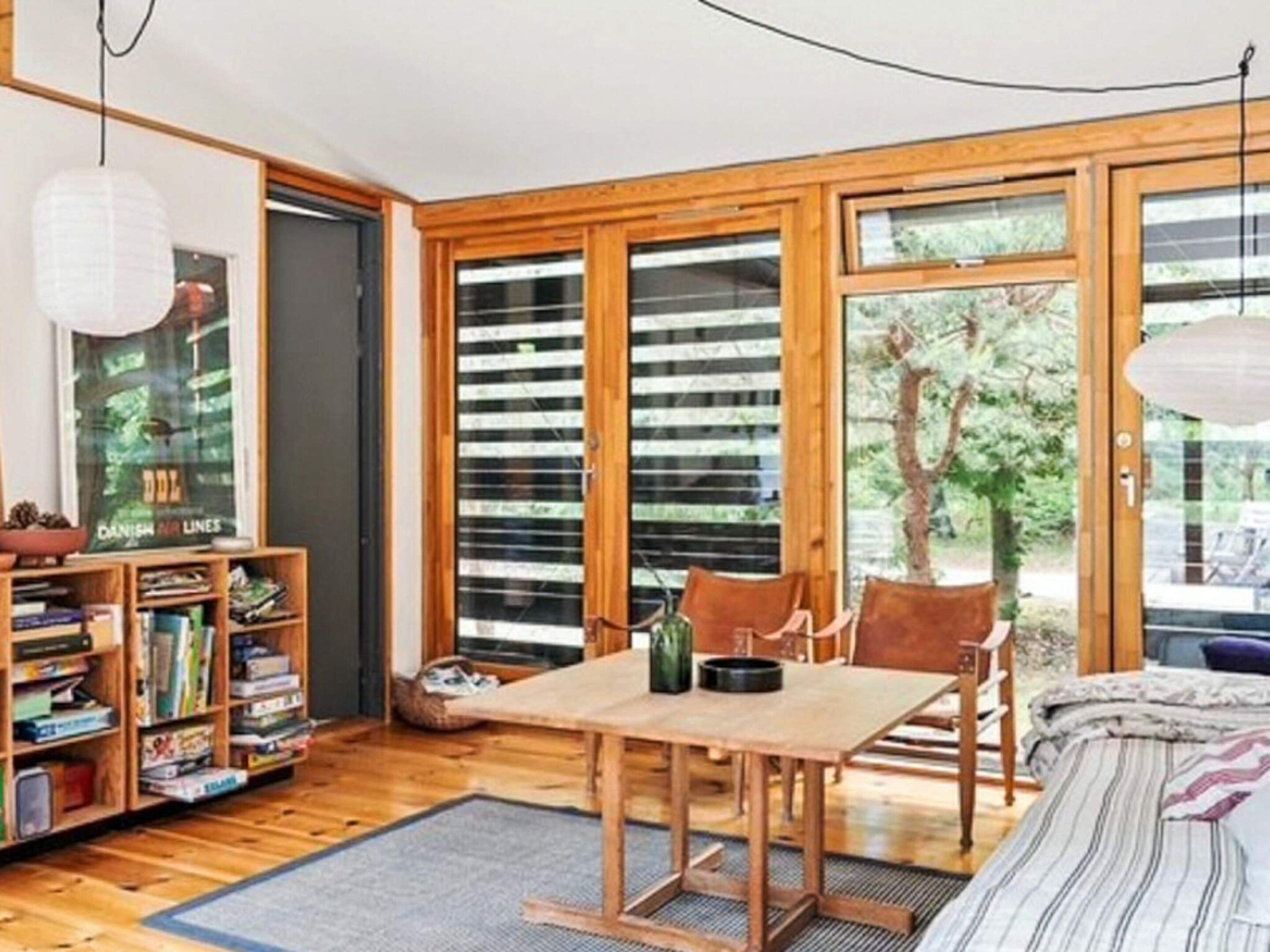 A room with a wooden floor, a bed with a striped blanket, a table with chairs, a bookshelf, and large windows with blinds.