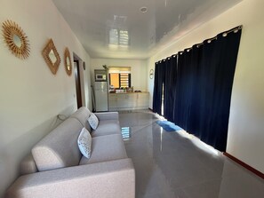 A living room with a sofa, a kitchen in the background, and a hallway leading to a bedroom.