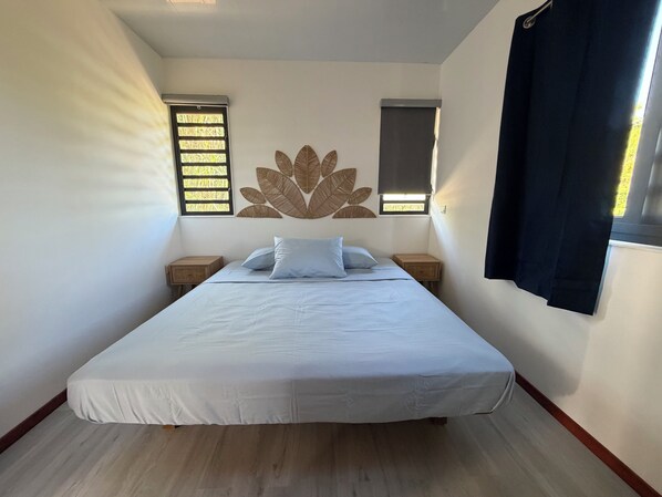 A bedroom with a bed, two nightstands, a wall-mounted leaf decoration, and two windows with blinds.