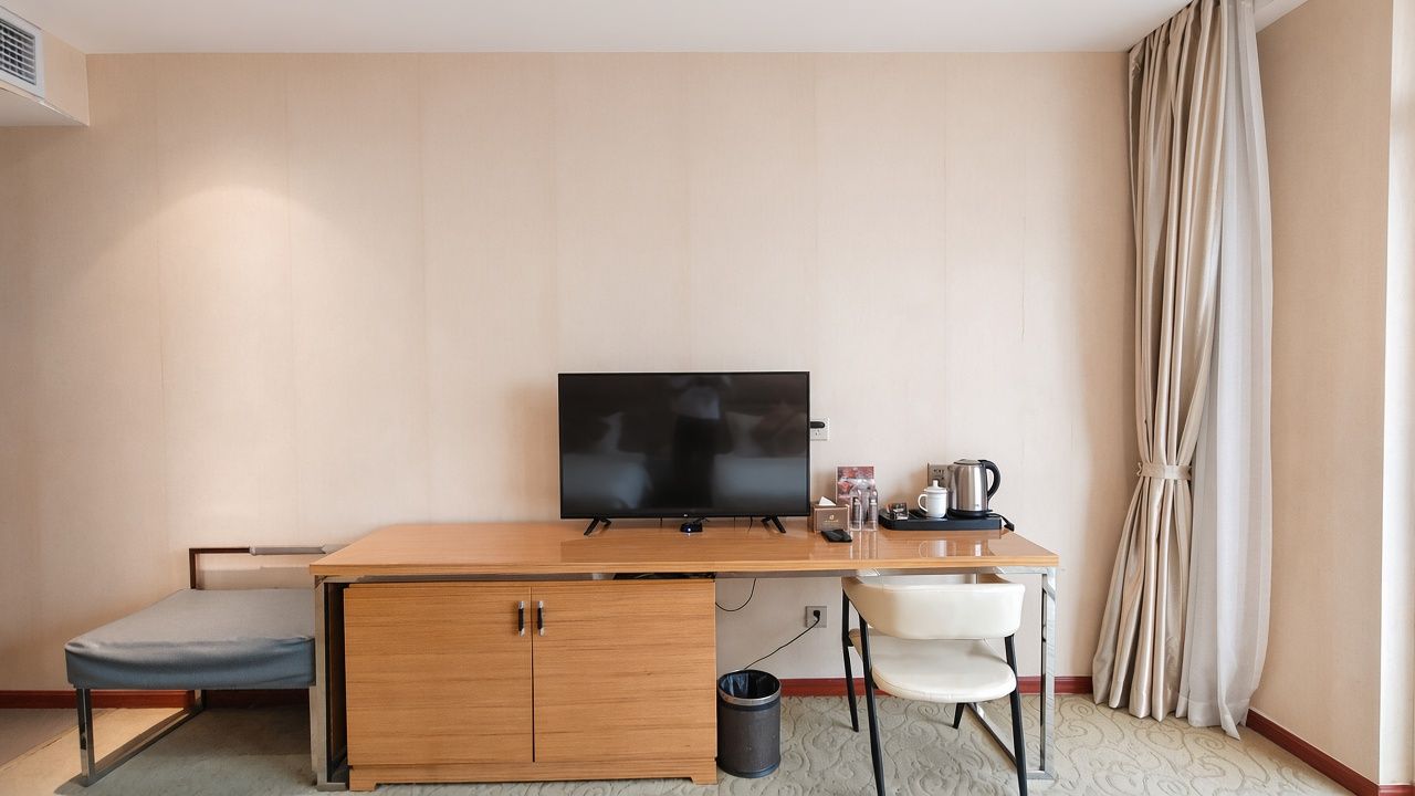 Une chambre d’hôtel compacte comprenant un lit, un bureau avec une télévision, une chaise et une petite table avec une bouilloire et des tasses.