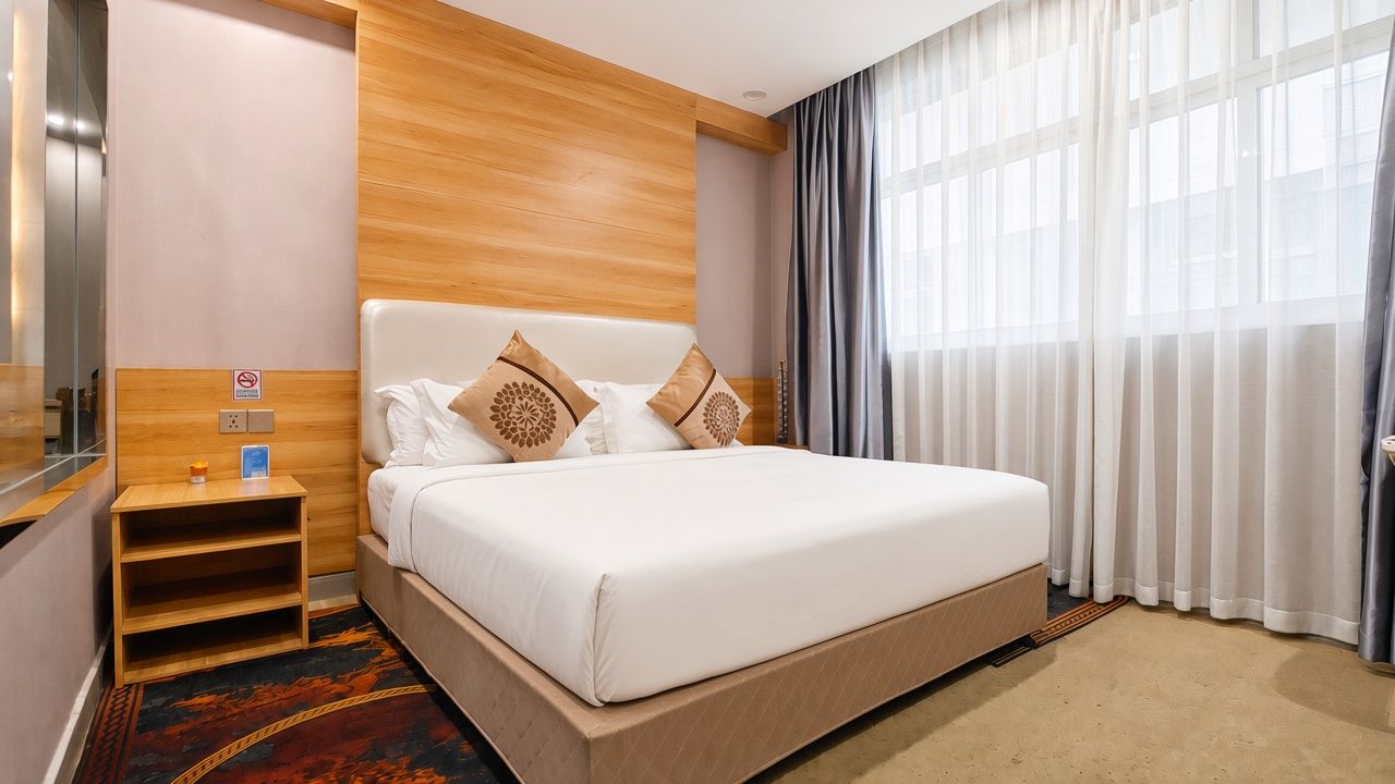 A hotel room with a bed, a nightstand, a wooden headboard, a window with curtains, and a carpet.