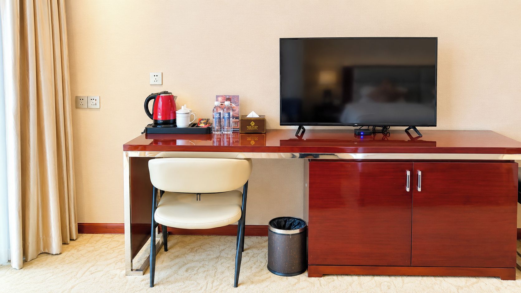 A hotel room with a desk, a chair, a television, a trash can, and a power outlet.