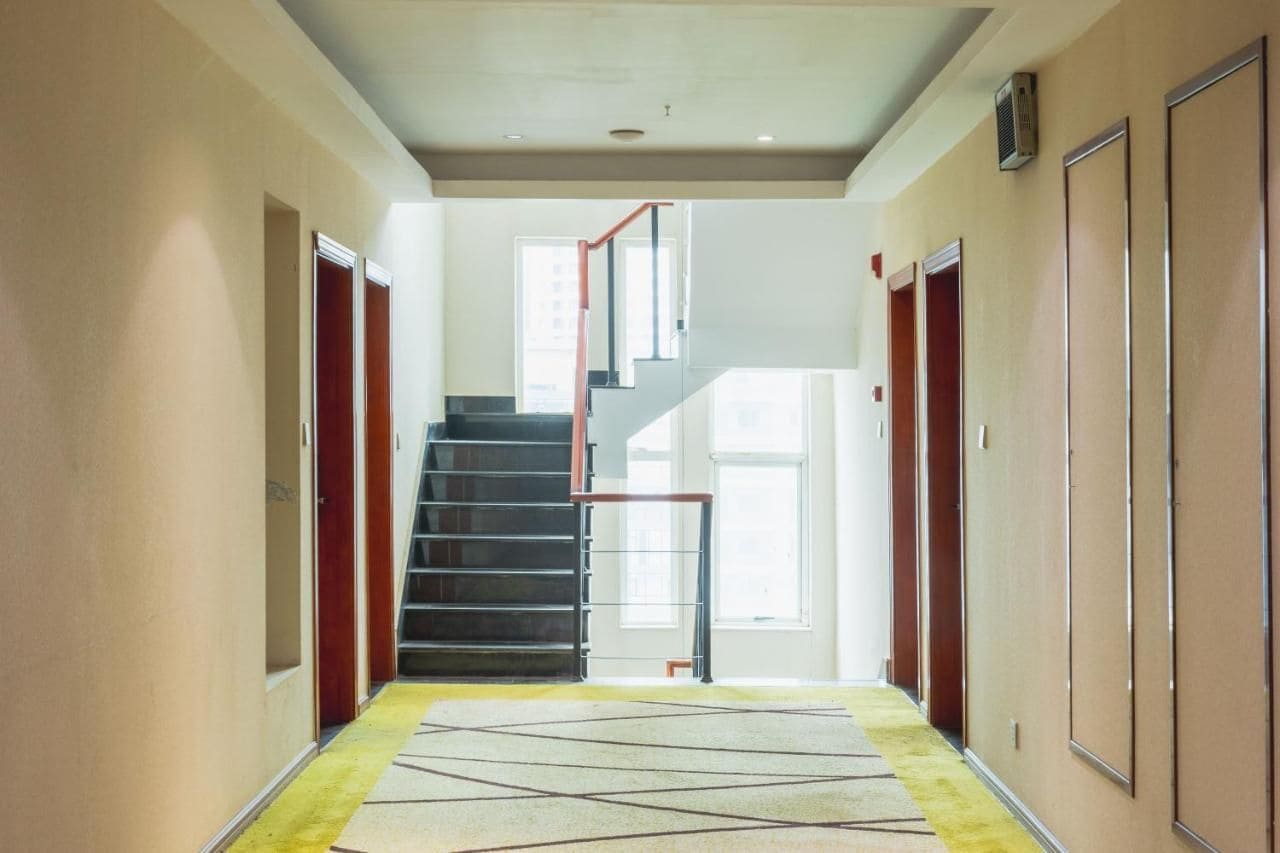 A hotel hallway with a central staircase, doors on either side, and a patterned carpet.