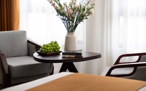 Una habitación con un sofá gris, una mesa redonda con un florero y un bol de uvas verdes, y una pila de libros.