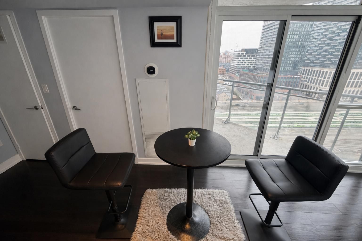 Una habitación moderna con dos sofás de cuero negro, una mesita redonda con una planta y vistas a un paisaje urbano a través de amplios ventanales.
