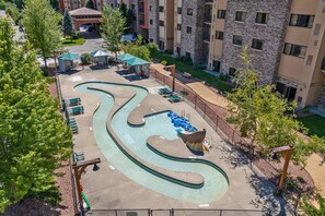 A swimming pool with a winding design, surrounded by lounge chairs and umbrellas, with a multi-story building in the background. - Dells Retreat - Sleeps 8 + Jacuzzi (Wisconsin Dells)
