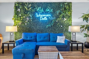 A room with a blue sectional sofa, two side tables with lamps, a wooden coffee table, and a green wall with a neon sign reading 'Wisconsin Dells'. - Dells Retreat - Sleeps 8 + Jacuzzi (Wisconsin Dells)