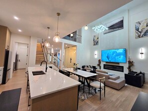 A modern kitchen with a central island, a dining area with a table and chairs, a living room with a sofa and a TV, and a staircase leading to an upper level.