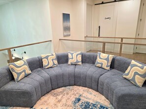 A curved gray sofa with patterned cushions, a round rug, and a minimalist decor with a wall-mounted artwork and a door labeled 'Laundry'.