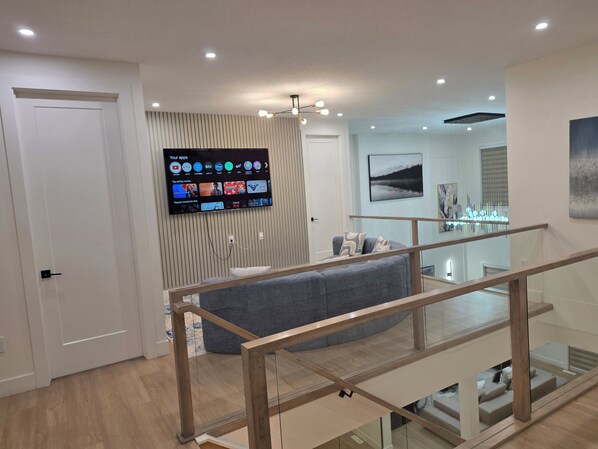 A modern living room with a flat-screen TV mounted on the wall, a grey sofa, and a staircase with glass railings.