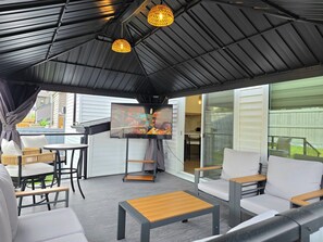 An outdoor patio with a covered seating area, a television screen, and a view of the backyard.