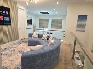 A modern living room with a curved grey sofa, a flat-screen TV mounted on the wall, and a staircase leading to an upper level.