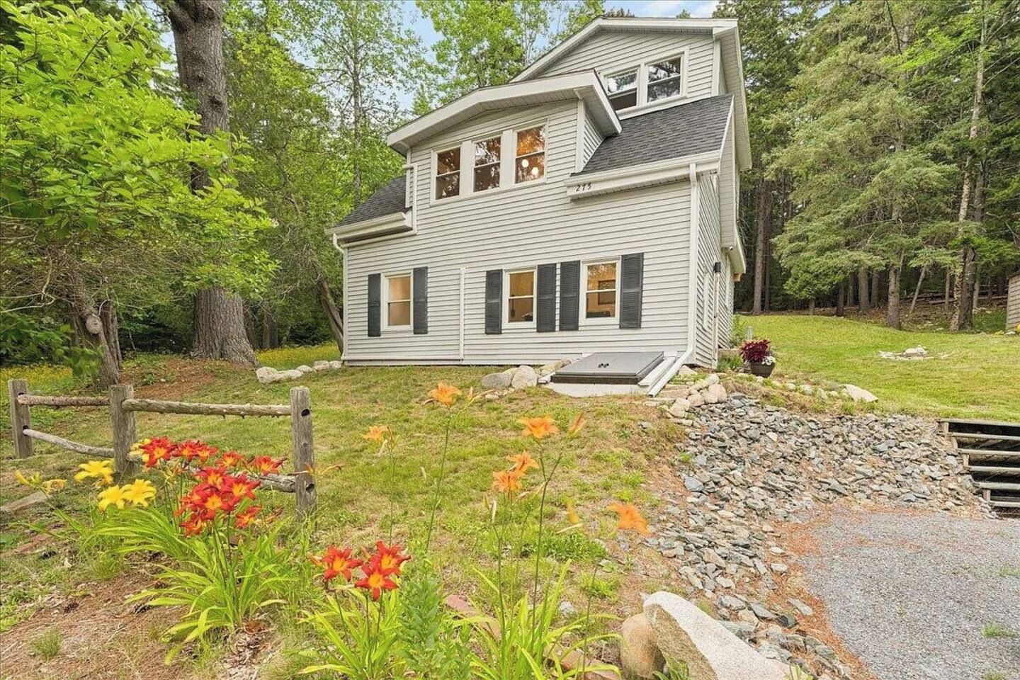 Une maison à deux étages avec un porche à l’avant, entourée d’un paysage naturel composé d’arbres et de plantes à fleurs.