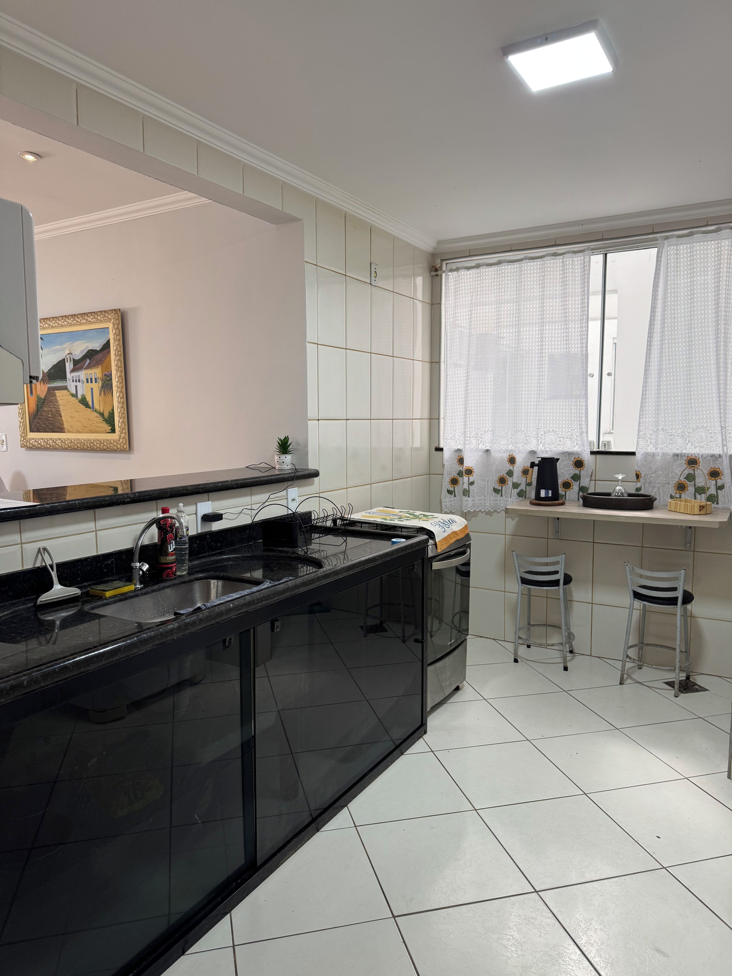 Una cocina con encimera negra, azulejos blancos, un cuadro y una ventana con cortinas.