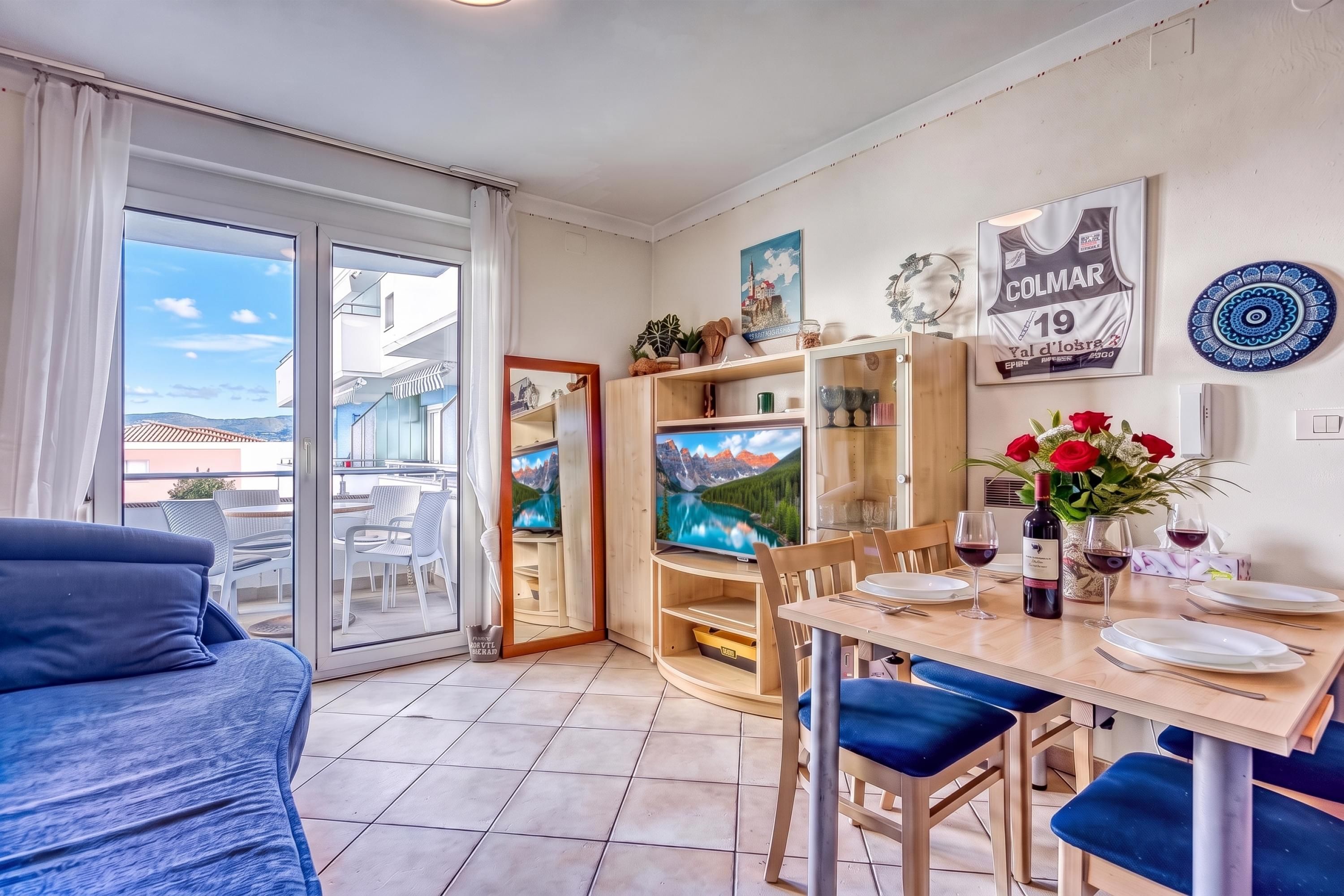 A modern living room with a sofa, dining table, and chairs, a TV, and a balcony with a scenic view.