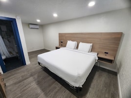 Habitación de hotel con cabecera de madera, cama grande con ropa de cama blanca, mesita de noche con lámpara y aire acondicionado de pared.