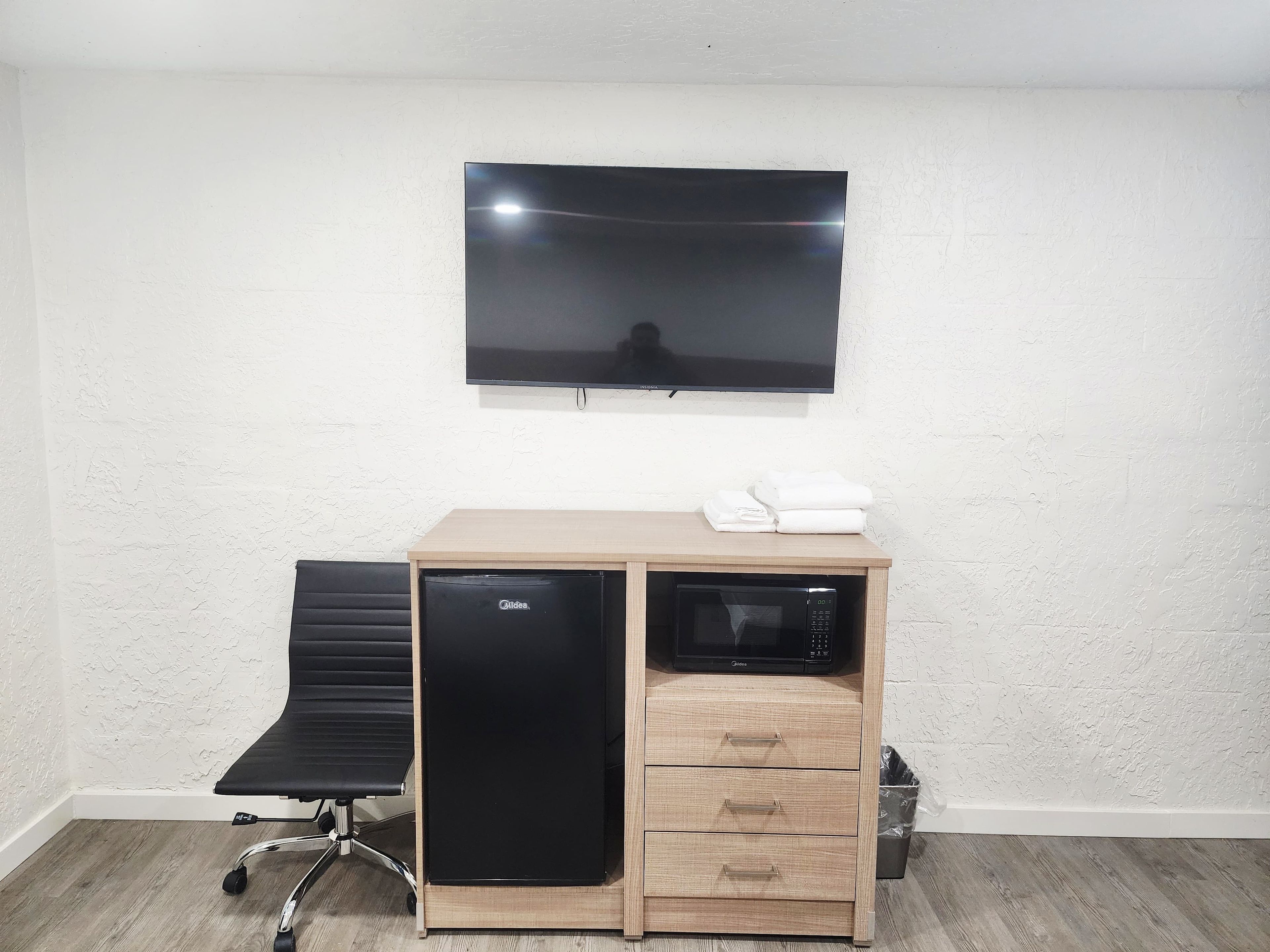 Habitación compacta con televisión de pantalla plana en la pared, una kitchenette pequeña con microondas y una silla de oficina negra.