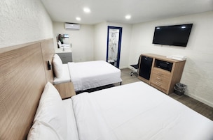 Habitación de hotel con dos camas, televisión, escritorio con silla, kitchenette pequeña y baño con ducha.