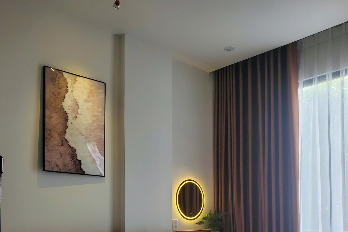 A hotel room with a large bed, a bedside table with a mirror, a painting on the wall, and a window with curtains.