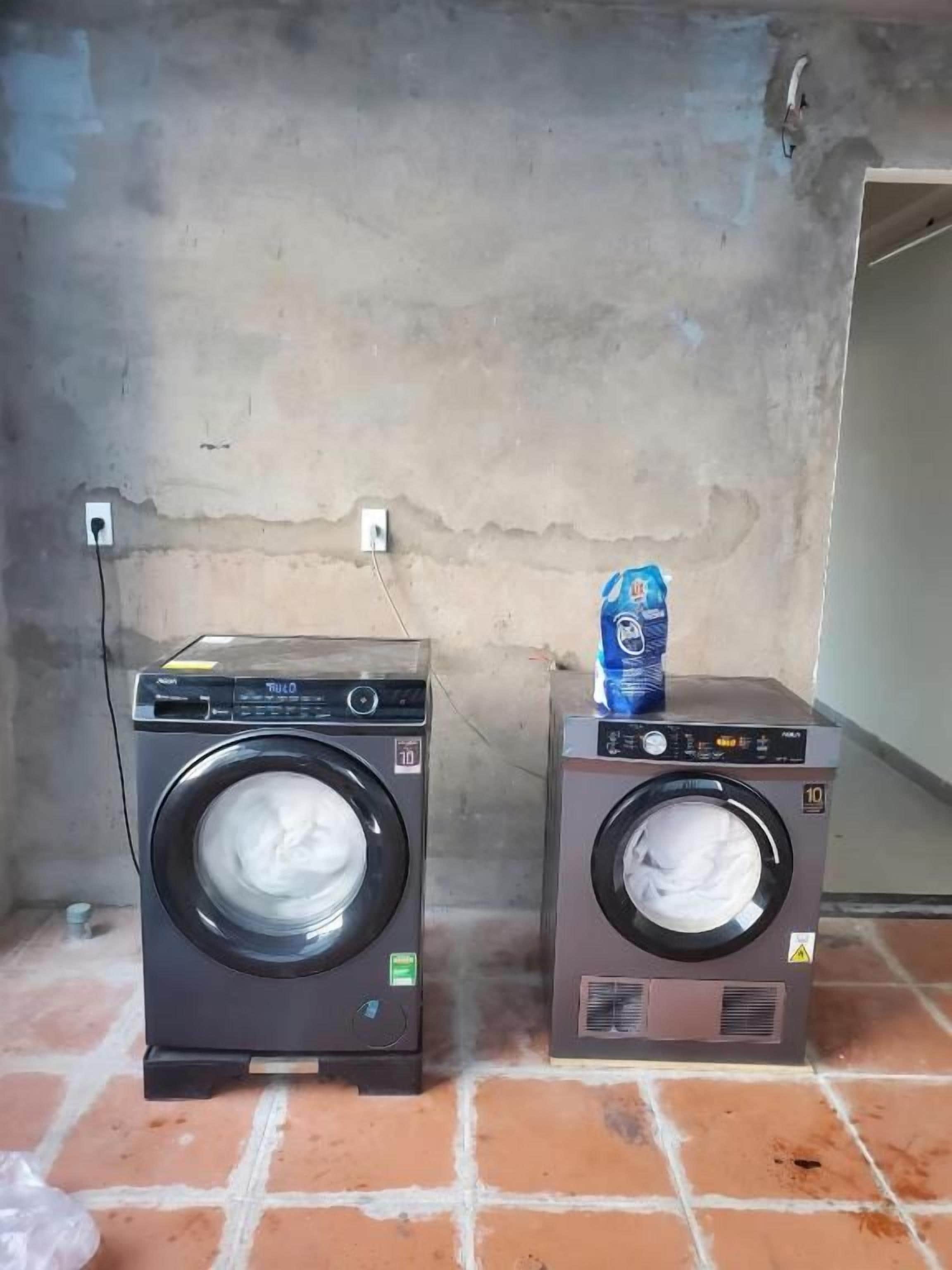 Dos lavadoras de carga frontal sobre un piso de baldosas, frente a una pared lisa con un enchufe.