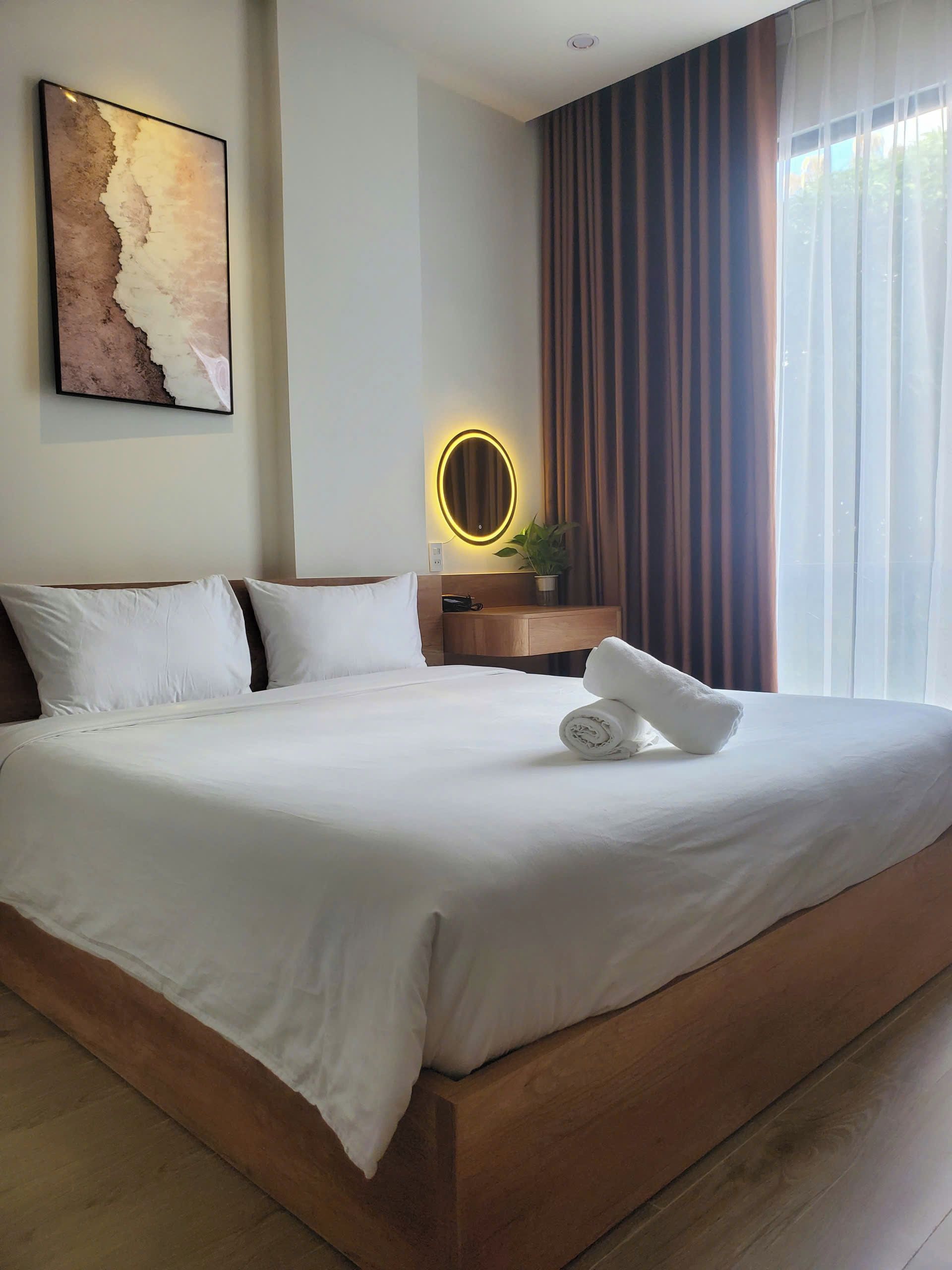 Una cama bien tendida con sábanas blancas, un cabecero de madera y una mesita de noche con un espejo.