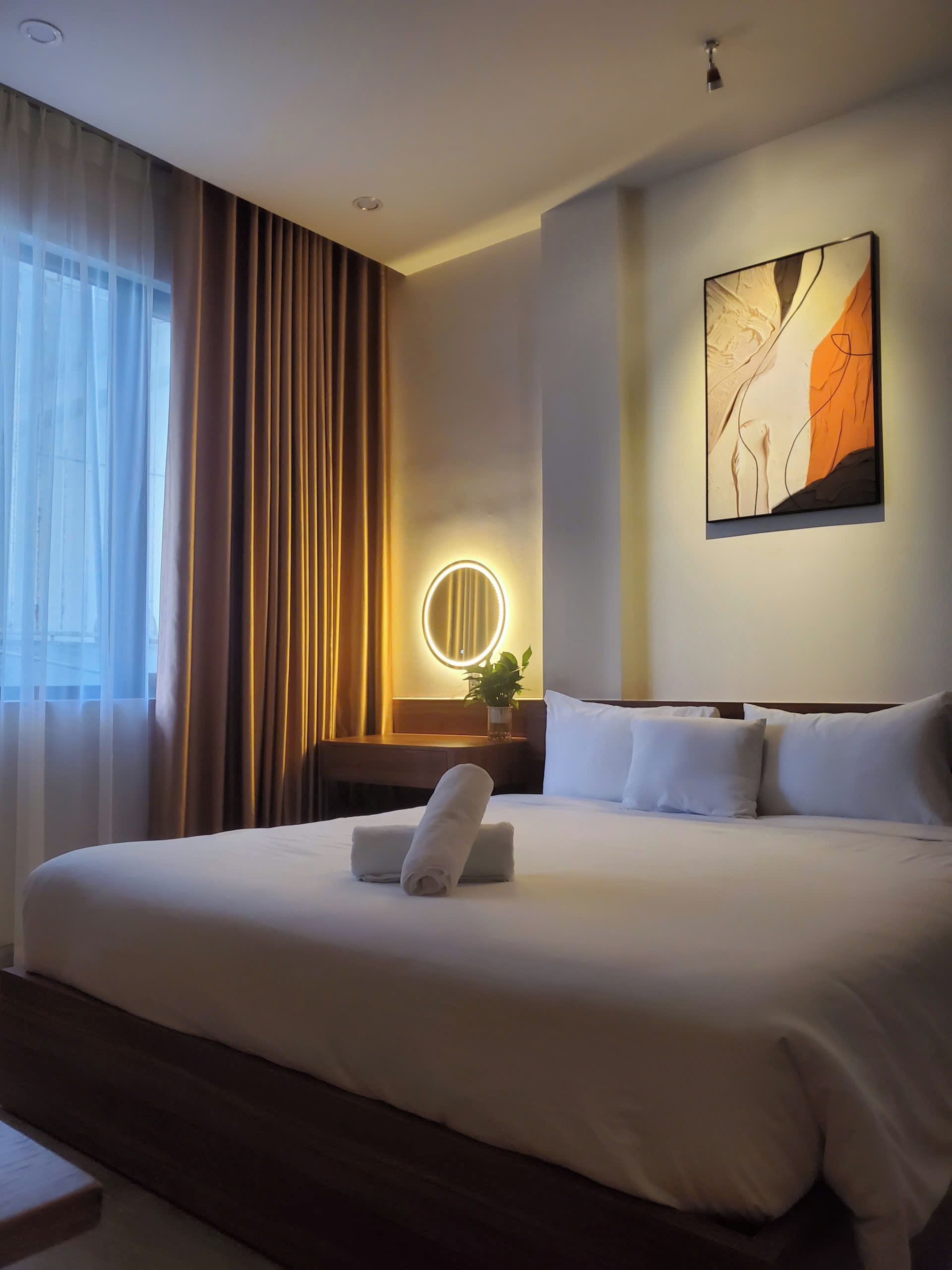 A hotel room with a large bed, a bedside table with a lamp, a painting on the wall, and a window with curtains.