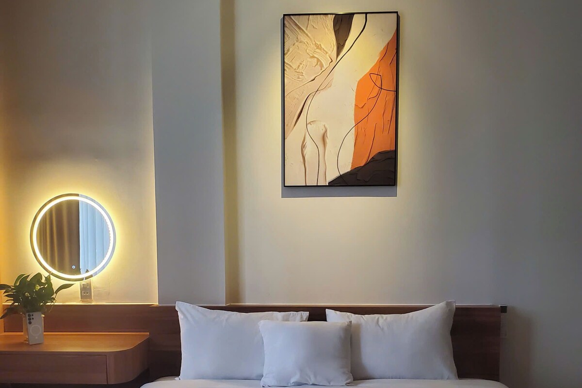 A hotel room with a bed, a bedside table with a lamp, a wall-mounted painting, and a round mirror.