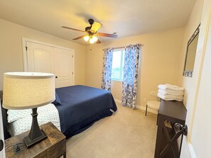 A bedroom with a bed, a ceiling fan, a window with curtains, a bedside table with a lamp, and a stack of towels.