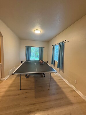 A room with a ping pong table, a window with curtains, and a ceiling light.