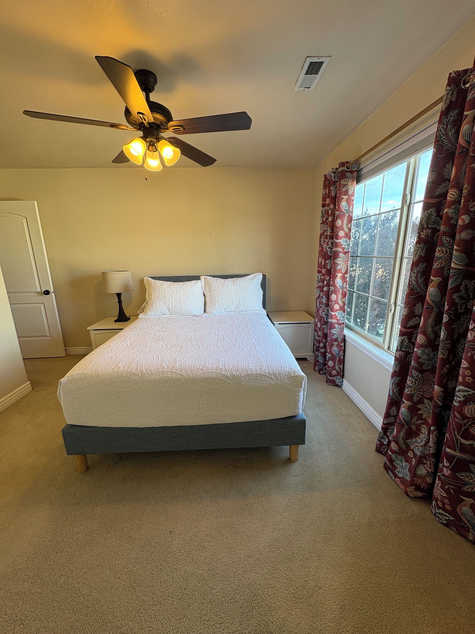 Quarto com cama, ventilador de teto, janela com cortinas e mesa de cabeceira com abajur.
