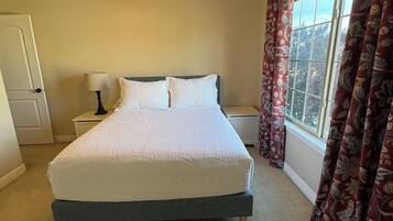 Quarto com cama, ventilador de teto, janela com cortinas e mesa de cabeceira com abajur.