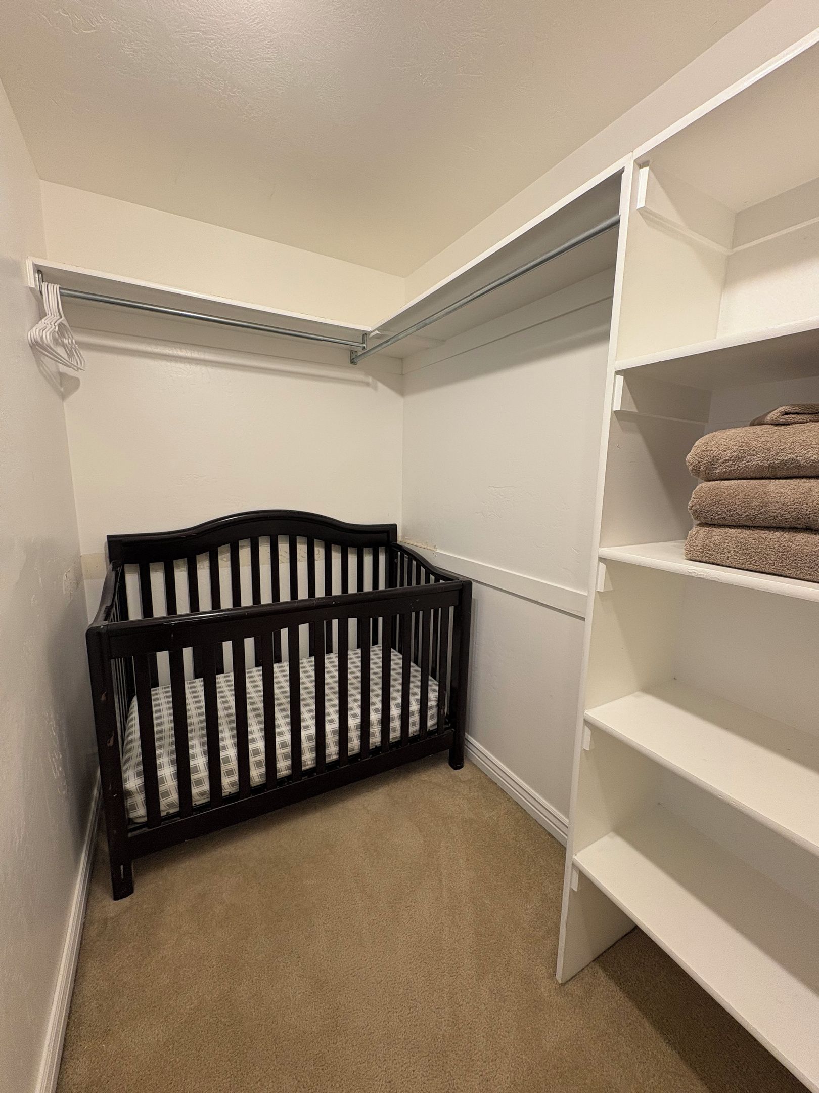 Quarto de canto com berço, prateleiras brancas e suporte para toalhas.