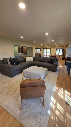 A spacious living room with a sectional sofa, a white ottoman, and a wicker chair. - 5-BR Family Retreat – Pool Table & Waffles (Provo)