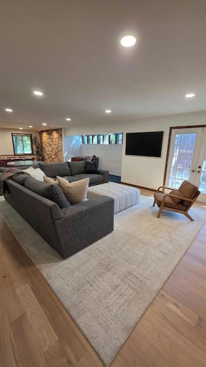 A modern living room with a sectional sofa, a wooden floor, a large window, and a flat-screen TV mounted on the wall.