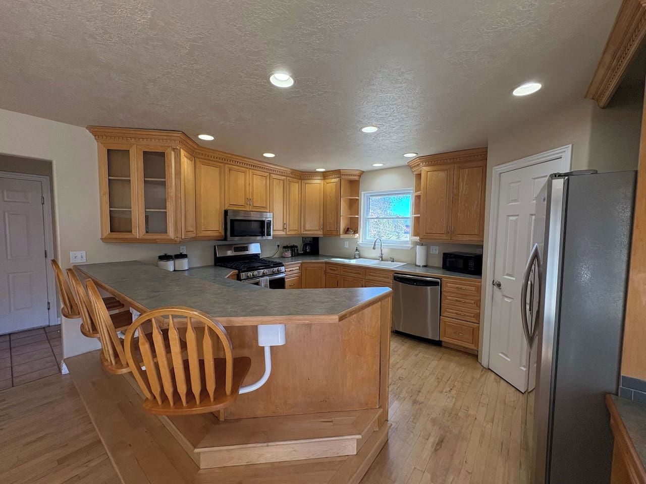 Una cocina moderna con armarios de madera, una isla central con asientos, electrodomésticos de acero inoxidable y un ventanal que proporciona luz natural.
