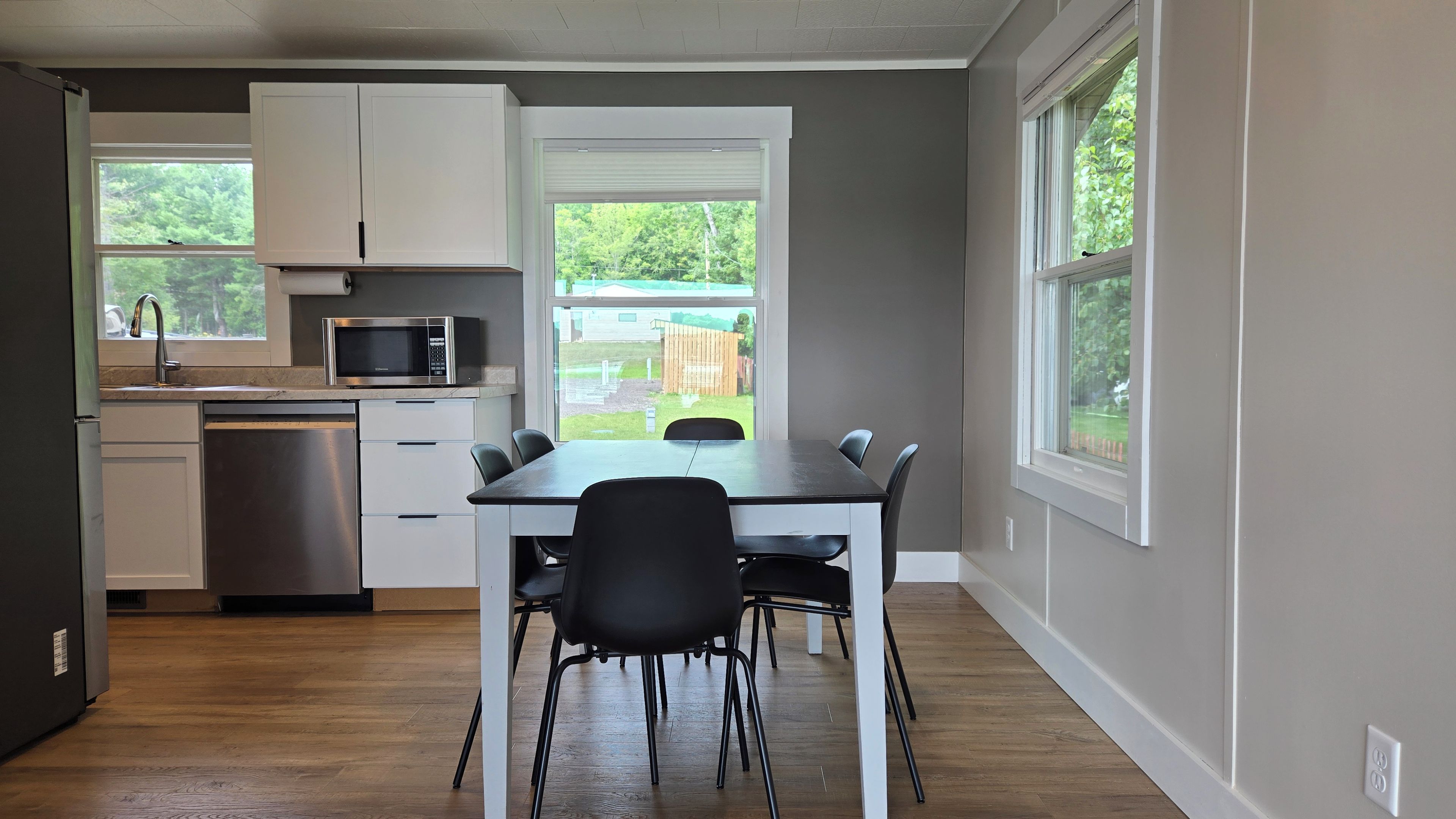A modern kitchen with white cabinets, a stainless steel microwave, and a dishwasher. There is a dining table with four chairs and a view of a backyard through a large window.